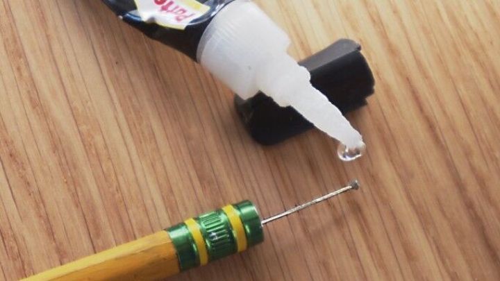 Eine Tube Klebstoff mit einem kleinen Tropfen auf einem Holztisch steht neben einem Bleistift mit einer feinen Nadelspitze.
