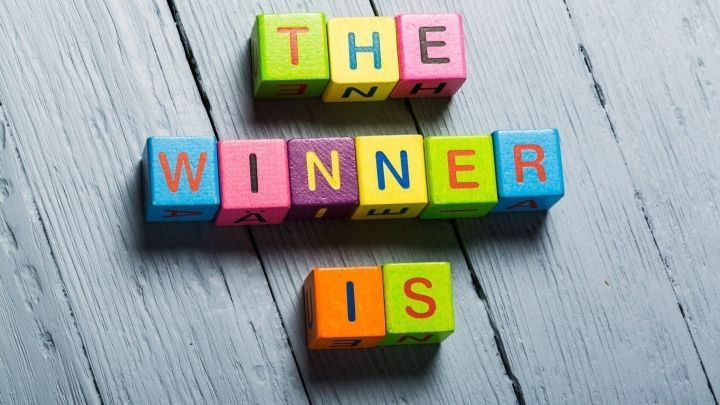 Bunte Buchstaben auf einem Holztisch bilden den Satz "THE WINNER IS", umgeben von einem neutralen Hintergrund.