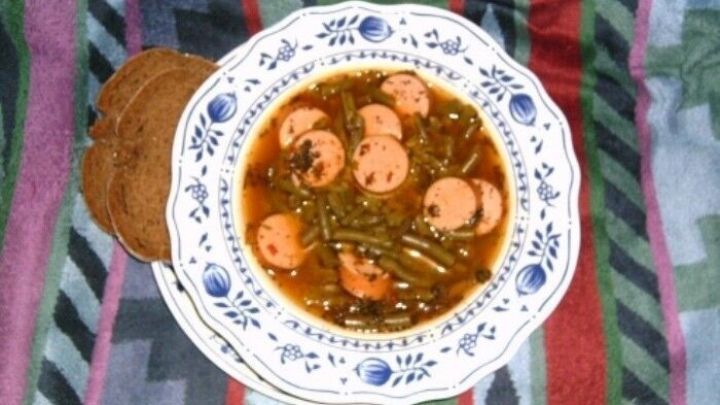 Eine Sch&uuml;ssel Bohnensuppe mit Bockwurstst&uuml;ckchen liegt auf einem bunten, gestreiften Tuch, umgeben von einem rustikalen Teller.