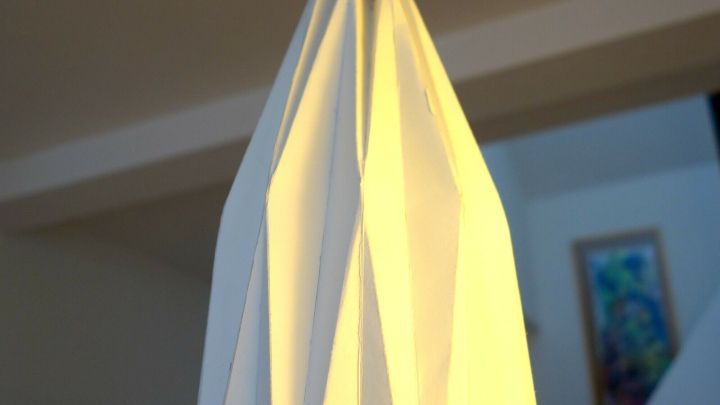 Ein eleganter Origami-Lampenschirm h&auml;ngt von einer blauen Schnur und strahlt warmes Licht in einem modernen Raum aus.