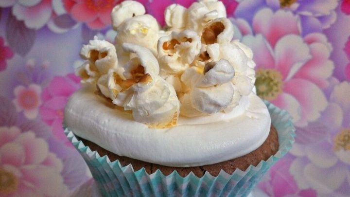 Ein Schoko-Cupcake mit cremigem Topping wird kunstvoll mit knusprigem Popcorn verziert, vor einem bunten Blumenhintergrund.