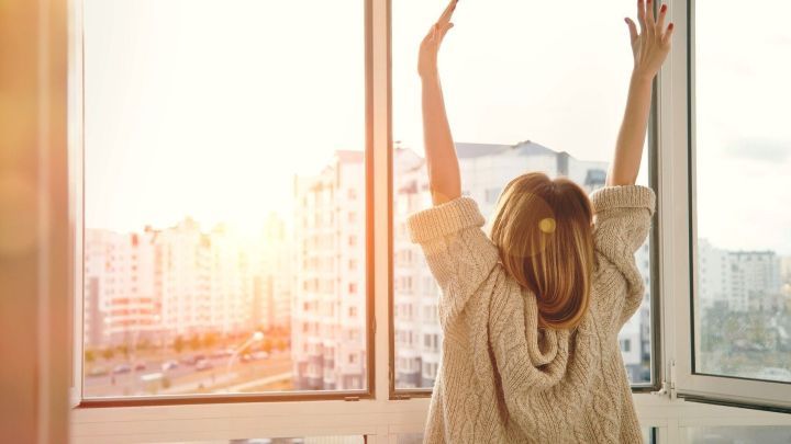 Eine Person steht vor einem Fenster, reckt die Arme nach oben und genie&szlig;t das Licht des Morgen, was ein Gef&uuml;hl von Wachheit vermittelt.