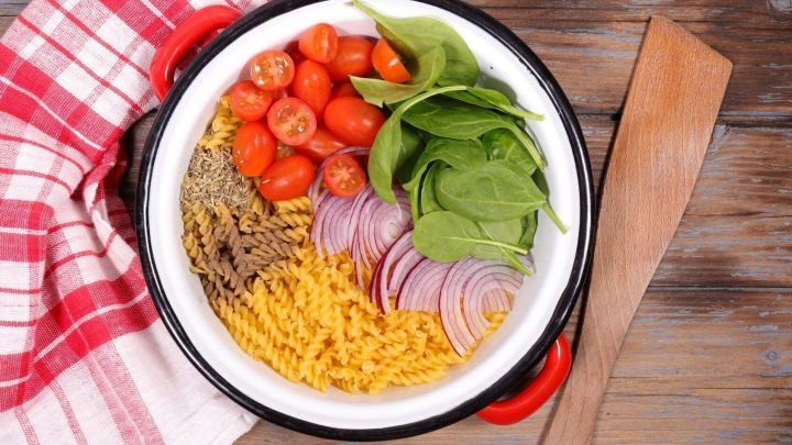 Eine bunte Mischung aus Tomaten, Spinat und Zwiebeln in einem Topf mit Pasta steht auf einem Holzuntergrund, umgeben von einem K&uuml;chentuch.
