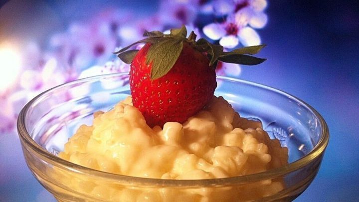 Eine Sch&uuml;ssel mit cremigem Milchreis, garniert mit einer Erdbeere, steht vor einem verschwommenen, bl&uuml;henden Hintergrund.