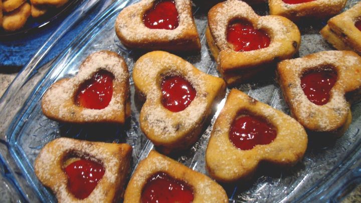 Herzpl&auml;tzchen aus Keksen mit Erdbeermarmelade in Herzform auf einer Glasschale, umgeben von anderen Pl&auml;tzchen.