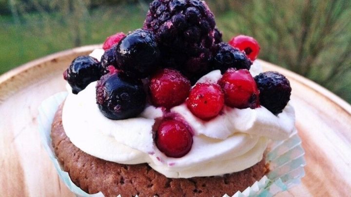Ein schokoladiger Muffin mit einer cremigen Schicht und bunten Beeren oben, auf einem Holzbrett im Freien platziert.