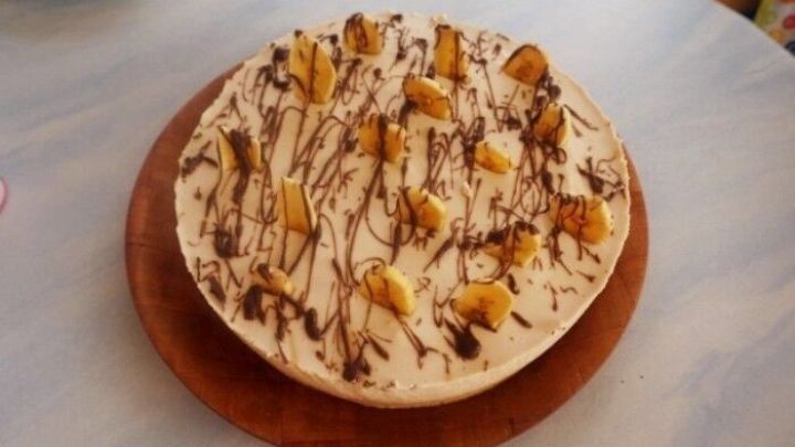 Eine Bananentorte liegt auf einem Holzbrett, dekoriert mit Schokolade und Bananenscheiben, umgeben von einem hellen Tisch.