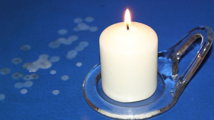 Eine brennende Kerze steht auf einem Glasuntersetzer, w&auml;hrend Wachsreste auf einer blauen Lacktischdecke liegen.