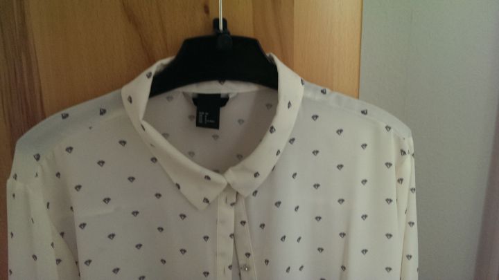 Ein wei&szlig;es Hemd mit d&uuml;nnem Kragen h&auml;ngt an einem schwarzen Kleiderb&uuml;gel vor einer Holzwand, bereit zum Spr&uuml;hen mit St&auml;rke.