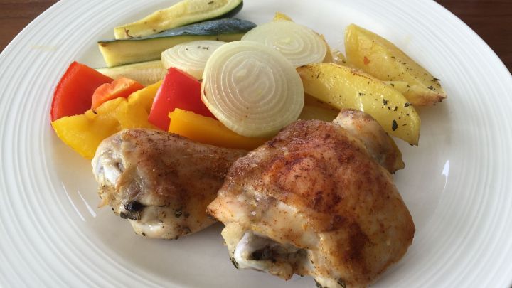 Gegrillte H&auml;hnchenschenkel liegen neben buntem Ofengem&uuml;se auf einem wei&szlig;en Teller, umgeben von einem schlichten Holzuntergrund.