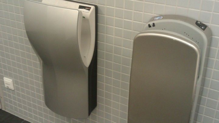 Ein H&auml;ndetrockner aus Edelstahl bl&auml;st Luft zur Trocknung von H&auml;nden in einem modernen, gefliesten Badezimmer.