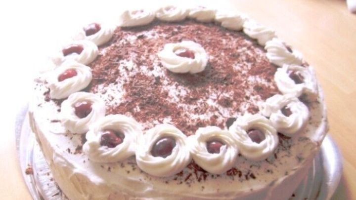 Eine Schwarzw&auml;lder Kirschtorte steht auf einem silbernen Teller, mit Sahne und Kirschen dekoriert, in einem hellen Raum.