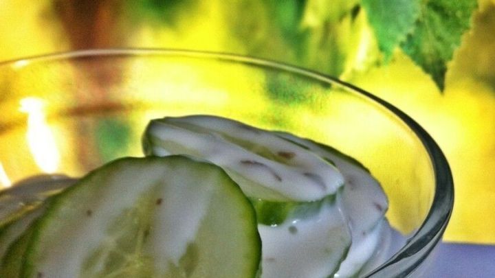 In einer Glassch&uuml;ssel liegt Gurkensalat mit Sauerrahm, umgeben von einem sanften, gr&uuml;nen Hintergrund.