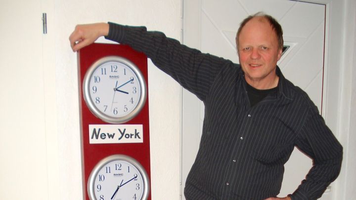 Ein Mann steht neben einer selbstgebauten Weltzeituhr mit drei Ziffernbl&auml;ttern f&uuml;r New York, Sydney und Capetown.