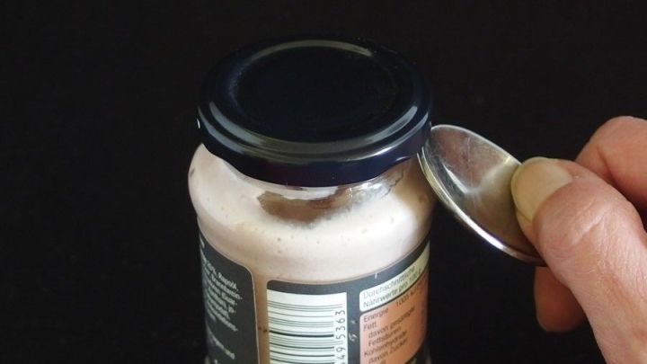 Ein Schraubglas wird mit einem L&ouml;ffel ge&ouml;ffnet, w&auml;hrend der Hintergrund schwarz ist. Das Objekt steht im Fokus.