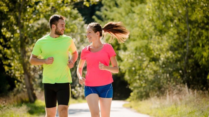Ein Mann und eine Frau joggen in bunten Sportshirts auf einem sonnigen Weg, umgeben von gr&uuml;ner Natur.