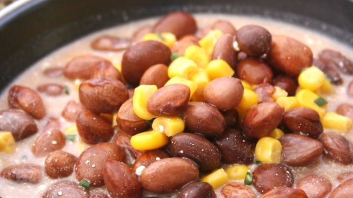 Eine bunte Bohnensuppe mit roten und gelben Bohnen sowie Mais f&uuml;llt eine Pfanne, umgeben von einer rustikalen Kulisse.
