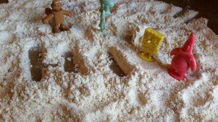 In einem sandigen Bereich sind bunte Spielsachen sichtbar, die im feinen Spielsand platziert sind, was ein kreatives Spielumfeld schafft.