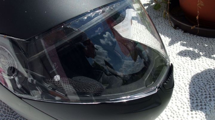 Ein schwarzer Motorradhelm mit klarer Visierfl&auml;che liegt auf einem hellen Untergrund, umgeben von Pflanzen und Sonnenlicht.