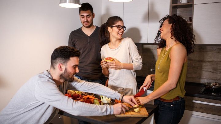 Eine Gruppe von vier Freunden bereitet gemeinsam ein Gericht in einer modernen K&uuml;che mit einem gro&szlig;en Holzbrett vor.