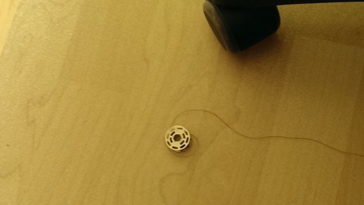 Ein Garnspulen-Objekt liegt auf dem Boden, w&auml;hrend ein Stuhlbein in der N&auml;he steht, und ein Faden zieht sich davon weg.