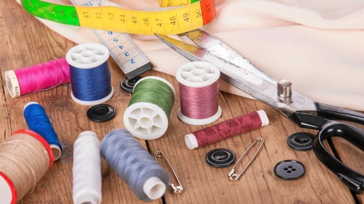 Ein Rollschneider liegt neben farbigen N&auml;hgarnen und Kn&ouml;pfen auf einem Holztisch, umgeben von N&auml;hzubeh&ouml;r.