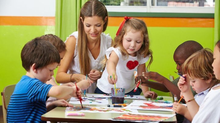 Sechs Kinder und eine Erzieherin malen gemeinsam an einem Tisch in einer kunterbunten Kita-Umgebung.