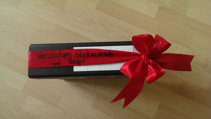 Ein schwarzer Ringordner mit rotem Geschenkband und einer Schleife liegt auf einem hellen Tisch.