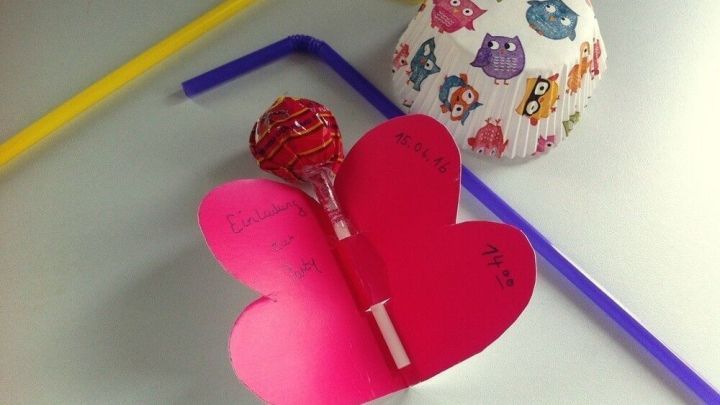 Eine pinke Einladungskarte in Schmetterlingsform h&auml;lt einen Lolli. Bunte Strohhalme und ein Pappteller sind im Hintergrund.