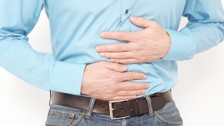 Ein Mann in einem blauen Hemd h&auml;lt sich den Bauch, was auf Sodbrennen hindeutet, in einem neutralen Raum steht er da.