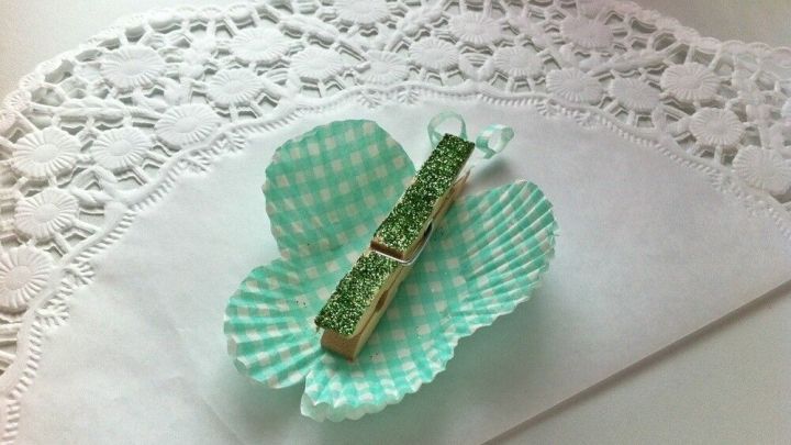 Eine gr&uuml;ne Muffinform ist dekorativ gefaltet und h&auml;lt eine Holzklammer, die als Schmetterling dient, auf einem wei&szlig;en Tisch.