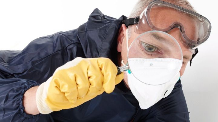 Eine Person untersucht mit einer Lupe Schimmel, tr&auml;gt dabei Schutzausr&uuml;stung und ist in einem hellen Raum aktiv.