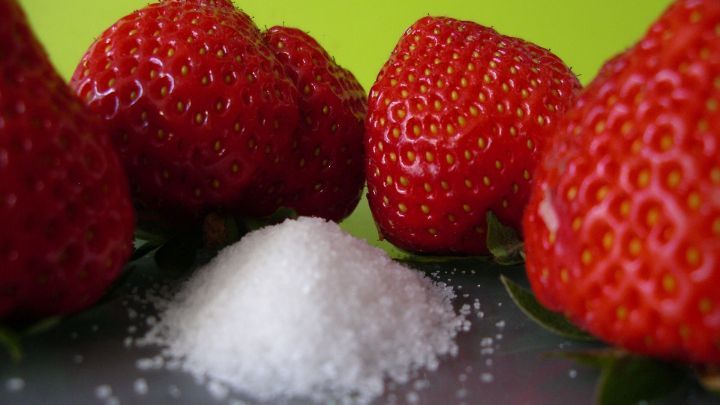Frische Erdbeeren liegen neben einer kleinen Menge Zucker, auf einer farbigen Unterlage, die Fruchtzucker symbolisiert.