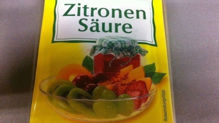 Eine Packung Zitronens&auml;ure steht auf einem Tisch, umgeben von frischen Fr&uuml;chten, die ihre vielseitigen Einsatzm&ouml;glichkeiten zeigen.