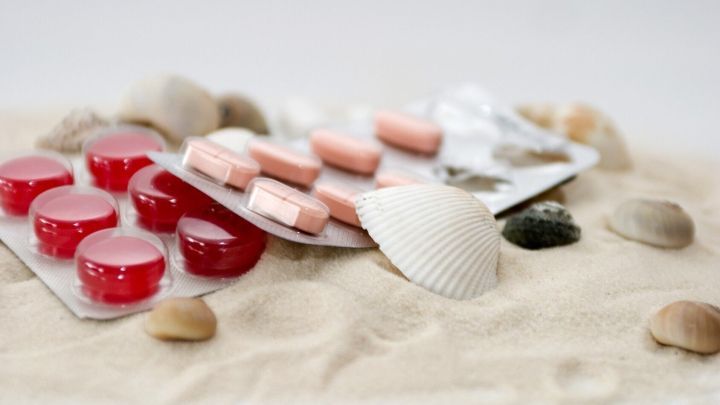 Rote und rosa Tabletten liegen auf Sand, umgeben von Muscheln, was den Kontext eines Urlaubs darstellt und auf eine Reiseapotheke hinweist.
