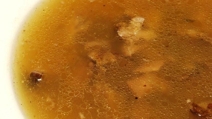 Eine br&auml;unliche Br&uuml;he in einer wei&szlig;en Sch&uuml;ssel, sichtbar mit kleinen Fleischst&uuml;cken, im kulinarischen Kontext.