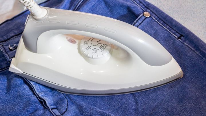 Ein B&uuml;geleisen liegt auf einer blauen Jeans, w&auml;hrend es versucht, Kaugummi zu entfernen. Es befindet sich auf einem neutralen Untergrund.