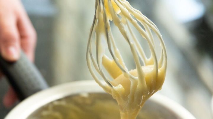 Ein R&uuml;hrbesen zieht cremigen R&uuml;hrteig aus einer metallenen Sch&uuml;ssel, w&auml;hrend im Hintergrund eine moderne K&uuml;che zu sehen ist.