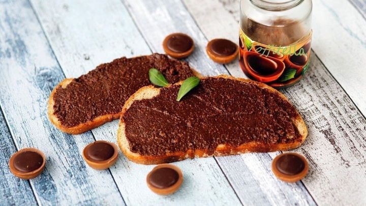 Zwei Brotscheiben mit schokoladigem Toffitella-Aufstrich liegen auf einem rustikalen Tisch, umgeben von Toffifees und einem Glas.