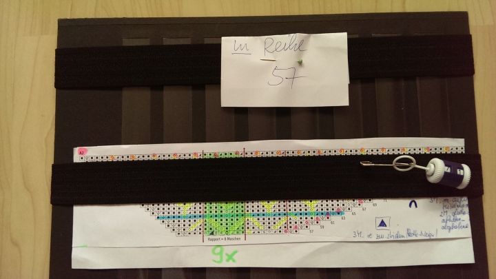 Ein Strickmusterblatt liegt auf einem Tisch, w&auml;hrend eine Maschenprobe mit farbigen Markierungen darauf sichtbar ist.