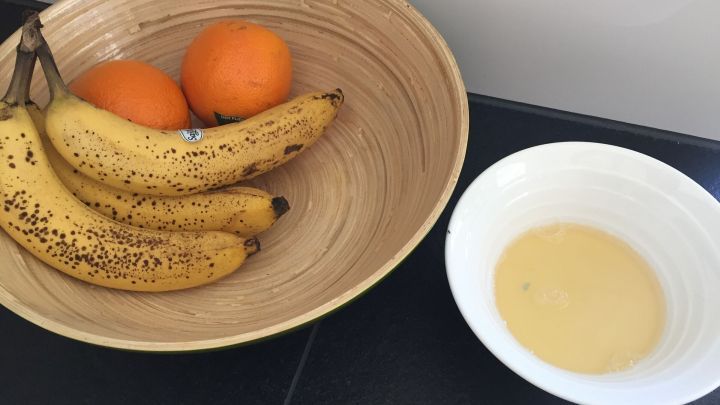 Reife Bananen und Orangen liegen in einer Schale, neben einer kleinen Sch&uuml;ssel mit einer gelben Fl&uuml;ssigkeit, die zum Fruchtfliegen-Fallen gedacht ist.