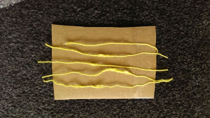 Gelbe Flusen liegen auf dem Seidengarn, das auf einem braunen Kartonst&uuml;ck pr&auml;sentiert wird, auf einem dunklen Untergrund.