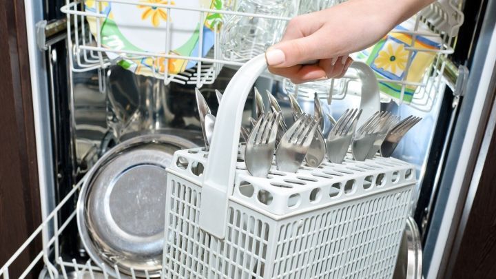 Ein Besteckkorb wird aus einem Geschirrsp&uuml;ler herausgehoben, um das Besteck zu entnehmen. Geschirr und Gl&auml;ser sind ebenfalls sichtbar.