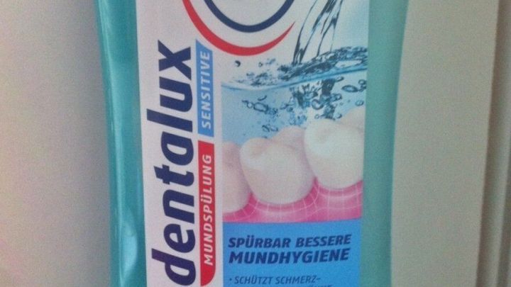 Eine Flasche Zahnsp&uuml;lung mit der Aufschrift &bdquo;dentalux Sensitiv&ldquo; steht auf einem hellen Hintergrund, umgeben von einer Wasserillustration.