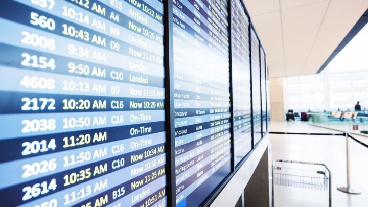 An einem Flughafen zeigen gro&szlig;e Anzeigetafeln Flugzeiten, einige sind versp&auml;tet, was Reisende verunsichert.