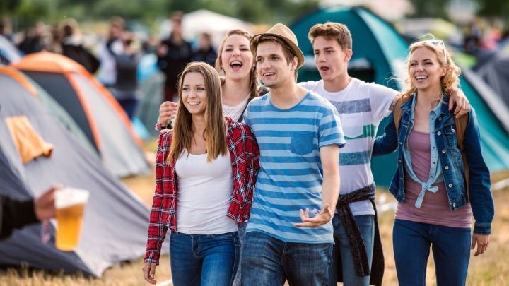 Eine Gruppe junger Menschen geht fr&ouml;hlich &uuml;ber ein Open-Air-Festival, umgeben von Zelten und anderen Besuchern.
