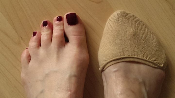 Ein nackter Fu&szlig; mit roten N&auml;geln steht neben einem beige F&uuml;&szlig;ling, der nur den Zehenbereich bedeckt, auf einem Holzfu&szlig;boden.