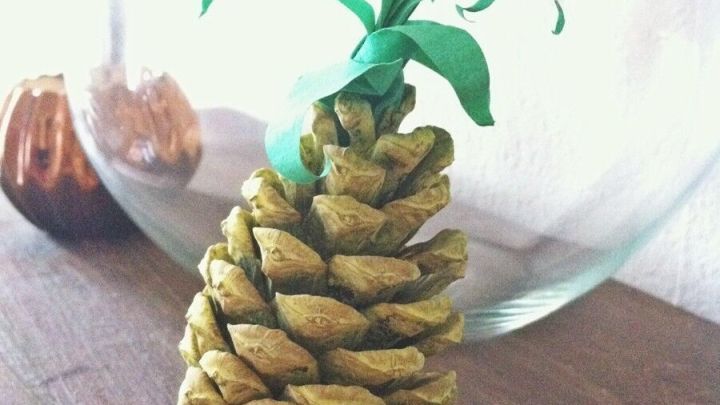 Ein Kiefernzapfen wurde kreativ zu einer Ananas mit gr&uuml;nen Bl&auml;ttern gestaltet, auf einem Holztisch vor einer Glasvase.