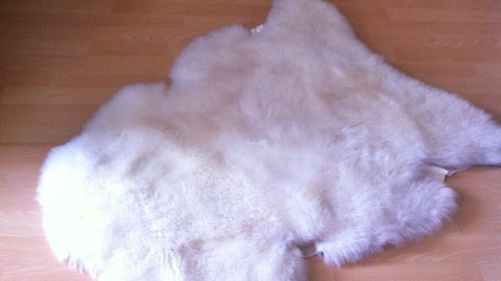 Ein weiches, helles Lammfell liegt auf einem Holzboden in einem gem&uuml;tlichen Raum, umgeben von Kerzen und einer Laterne.