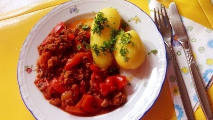 Ein Teller mit Paprika-Hackfleisch und Kartoffeln steht auf einem gelben Tischdecken, umgeben von Gl&auml;sern und Besteck.